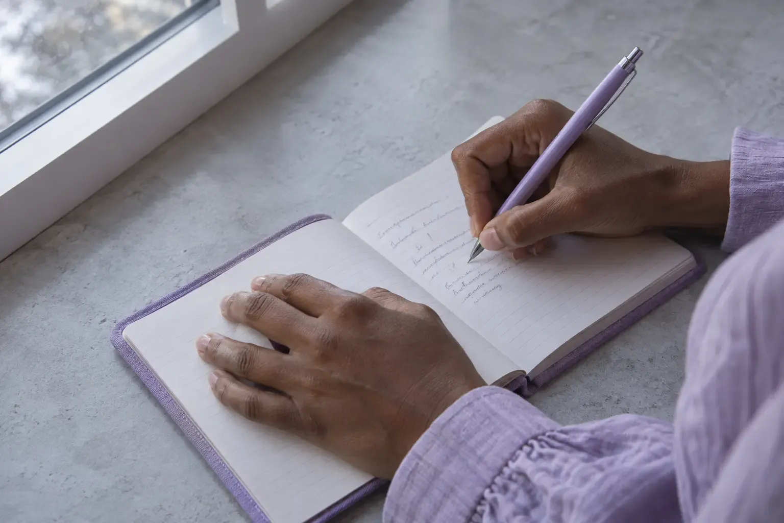 Close-up of hands writing in a lilac notebook beside a window.
