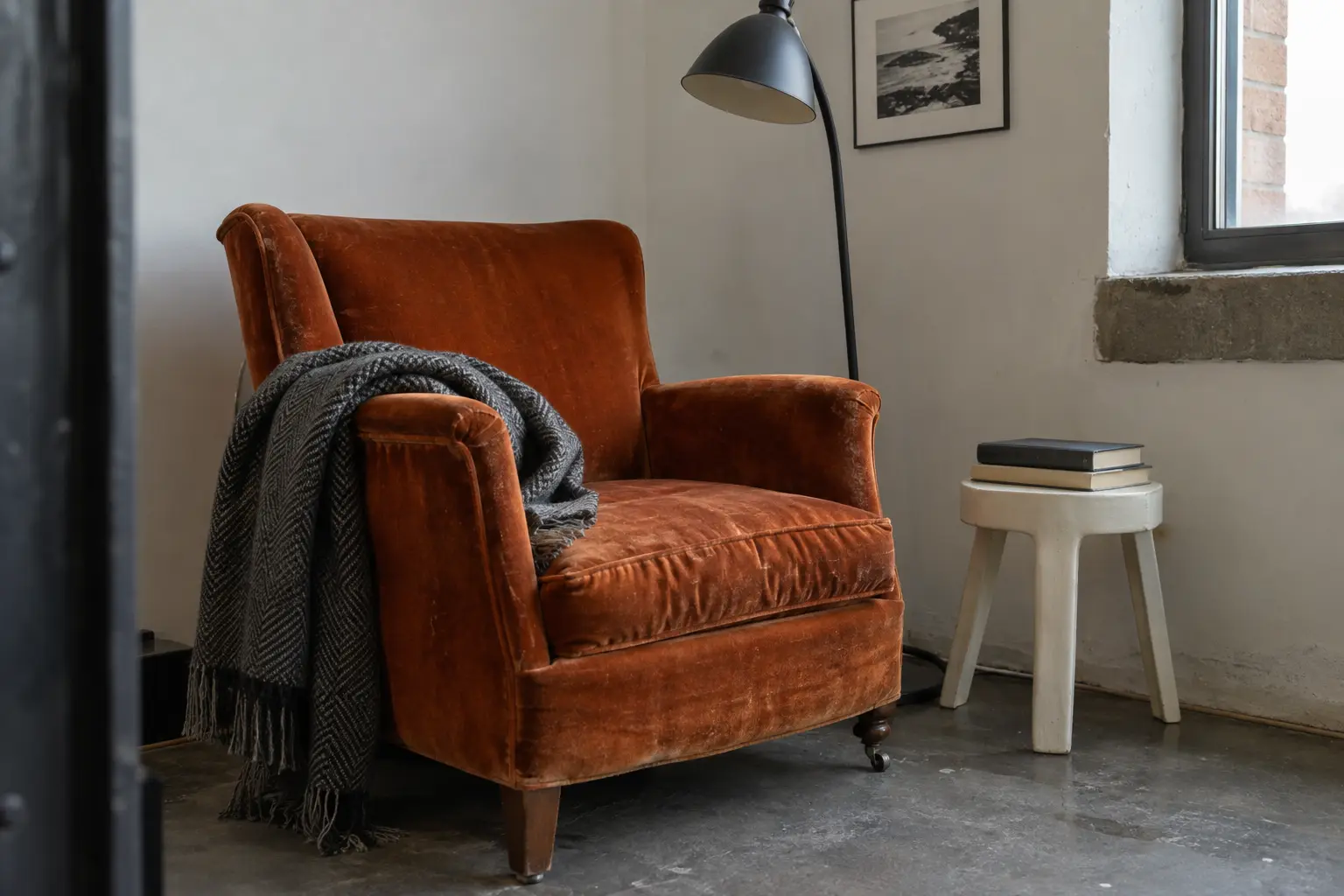 Rust-orange armchair with a charcoal throw, a black reading lamp, and books on a small white stool.