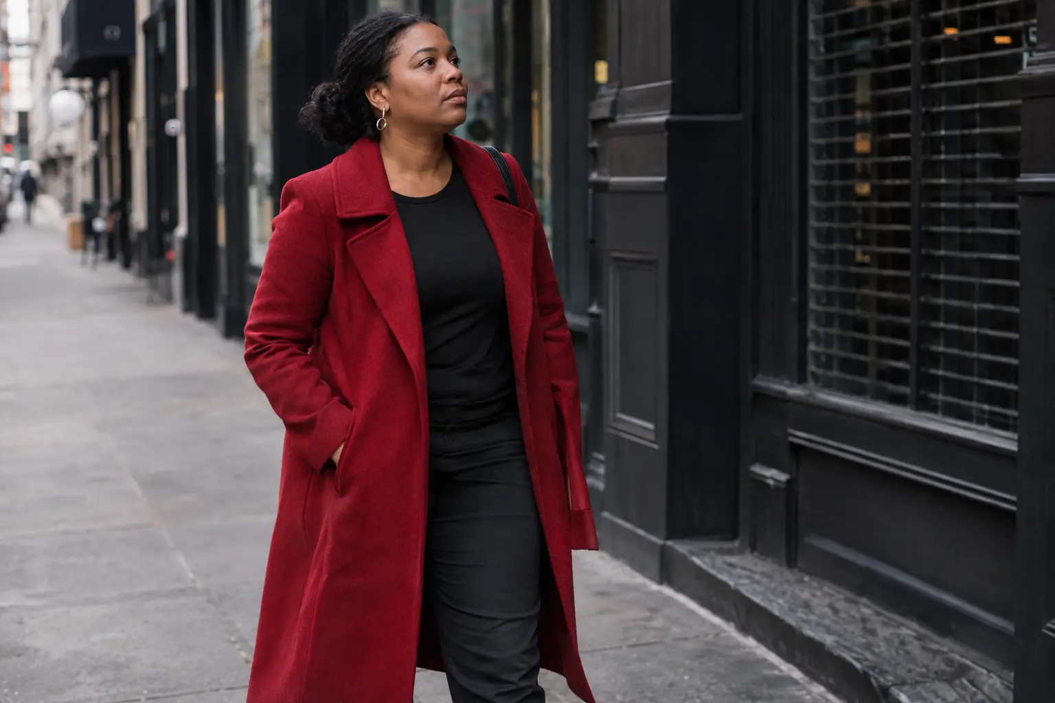 Woman in a crimson coat walking along a city sidewalk with her hands in her pockets.