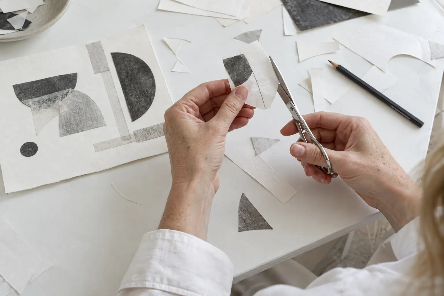 Hands cutting paper shapes for a collage on a white table with paper scraps and a pencil.